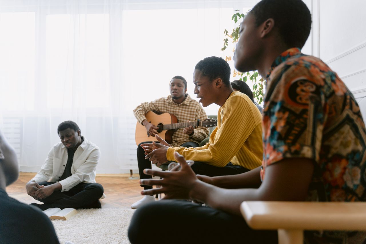Group of adults engaged in a musical session indoors, featuring singing and guitar playing.